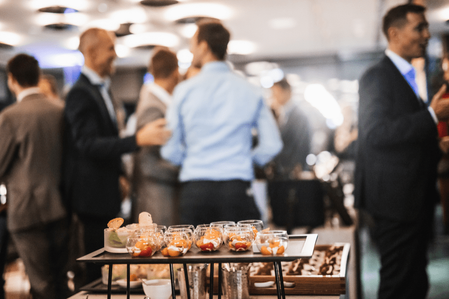 A professional catering display at a corporate event, featuring a selection of individual dessert or appetizer cups, some with fruit and others with pastries, arranged on a sleek black tray. The background shows a lively atmosphere with business professionals mingling and networking, highlighting Regency O'Fallon's catering services for corporate functions.