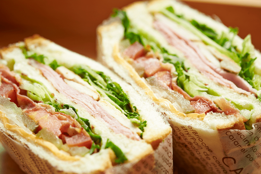 Close-up of two halves of a freshly made club sandwich, neatly wrapped and ready for catering 'to-go.' Layers of deli meat, crispy bacon, ripe tomatoes, crisp lettuce, and cheese are visible between slices of bread, perfect for Regency O'Fallon's corporate boxed lunch services