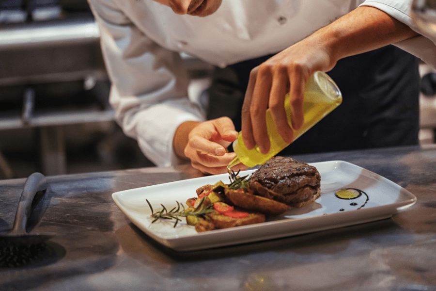 Close-up of a chef meticulously plating a gourmet meal, adding a final touch of olive oil to a juicy burger. This demonstrates the high-quality culinary experience provided by Regency O'Fallon for weddings, corporate events, and special occasions in O'Fallon, IL.